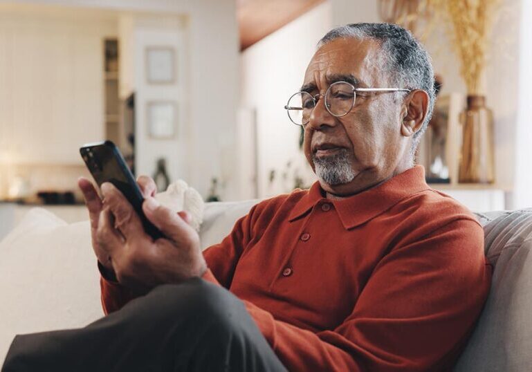 An older man sits on a living room couch, looking skeptically at his smartphone as he investigates a potential investment message, illustrating caution against online scams and financial fraud.