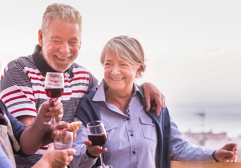 A smiling group of older adult friends socializing and toasting with glasses of red wine outdoors.